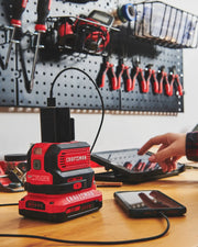 Photo of Craftsman CMCB1150B portable power station charging a smartphone via USB, on workbench with tools in background.