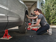 Person uses a CRAFTSMAN power tool to remove a car tire in driveway near garage. Tool set and jack stand visible.