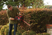 Photo of a person trimming hedges with a CRAFTSMAN cordless hedge trimmer, SKU CMCHTS820B, in a garden setting.