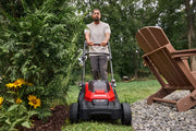 Photo of Craftsman lawn mower, SKU CMCMWP120M2, on grass next to plants and chair, operated outdoors.
