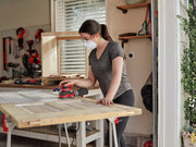 Person uses a CRAFTSMAN sander on wood, wearing a mask and goggles, in a workshop with tools visible in the background.