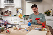 Person using Craftsman CMGL100B cordless glue gun for a solar system model craft on kitchen table with art supplies.