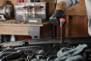 A person wearing black gloves is using a CRAFTSMAN T-handle hex key to work on a car engine. A set of T-handle hex keys is visible in the background on a workbench.