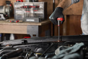 A person wearing a black glove uses a CRAFTSMAN T-handle screwdriver to work on a vehicle in a workshop. In the background, there is a set of CRAFTSMAN screwdrivers on a workbench.