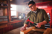 Photo of Craftsman red hammer CMHT51423 used to drive a nail into wood in a workshop setting.