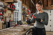 A person with a blurred face, wearing gloves, uses a red CRAFTSMAN hammer and a chisel to work on an old wooden door at a well-equipped workshop. Various tools and CRAFTSMAN products are visible on shelves and a pegboard in the background.