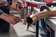 Photo of hands using a Craftsman ratchet wrench, SKU CMMT17560V, tightening a rusty bolt on a metal pipe in a workshop.