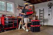 A person in a workshop holds a red and black CRAFTSMAN tool box. Two additional stackable CRAFTSMAN tool boxes are placed on the floor with a telescoping handle. The workspace contains a red tool chest, tools mounted on the wall, and a ladder leaning against the wall.