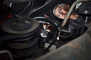 Close-up of a person using a ratchet wrench to work on a mechanical component inside a vehicle engine bay, surrounded by metal parts and pipes.