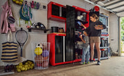 A garage space organized with CRAFTSMAN red and black storage cabinets and shelving. Various sports equipment and accessories are neatly stored, including balls in a wire basket, rackets, shoes, and bags. Two people are accessing items from the cabinets, one adult and one child, with the garage door partially open and natural light coming in.