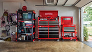 A garage interior with a red CRAFTSMAN tool chest and cabinet, both on wheels. The tool storage units feature multiple drawers for organizing tools and accessories. To the left is a black shelving unit with bins, boxes, and various household items, and sports equipment hanging on the wall.
