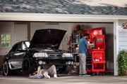 A garage scene showing a person working at a red CRAFTSMAN tool chest and cart, with another person lying under a black car performing repairs. The garage has shelves with various items and plants outside.
