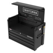 A black Craftsman tool chest with the top lid open, showing a work surface, pegboard back, power outlets, and three drawers below. The chest has the words 'CRAFTSMAN', '1776', 'USA', and '2026' printed on the back panel.