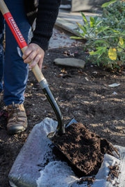 Photo of person using CRAFTSMAN shovel SKU CMXMLTP35329 to scoop soil from a bag in a garden setting.